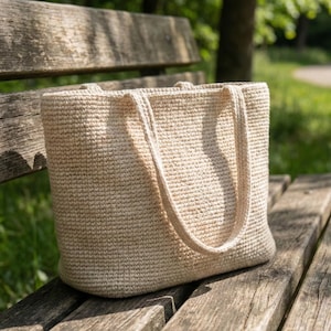 Könnte beinhalten: Eine beige, handgehäkelte Tragetasche mit stabilen Griffen, die auf einer verwitterten Holzparkbank liegt. Die strukturierte Oberfläche und die neutrale Farbe der Tasche deuten auf ein natürliches, umweltfreundliches Design hin, ideal für den Transport von Lebensmitteln oder persönlichen Gegenständen.
