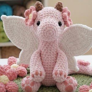 May include: A handmade pink crochet dragon with white wings, brown horns, and flower accents. The plush toy has black eyes and is sitting on a green surface. Additional pink crochet flowers are scattered around the dragon.