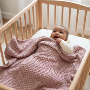 Pode incluir: Um bebé numa cama de madeira, envolto numa manta rosa de malha waffle. O bebé está a usar um body branco e a sorrir. A cama tem um colchão branco e ripas de madeira de cor clara.