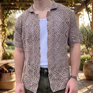 May include: A short-sleeved, taupe-colored crochet shirt featuring an open-knit pattern, a classic collar, and a front button-down closure. The garment is worn over a plain white tank top, showcasing the intricate, repeating square-based stitch design.
