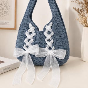 May include: A blue crochet handbag featuring two vertical lace-up sections on the front. Each section is threaded with white sheer ribbon that ties into a bow at the bottom. The bag has a single integrated handle and a textured, hand-stitched finish.