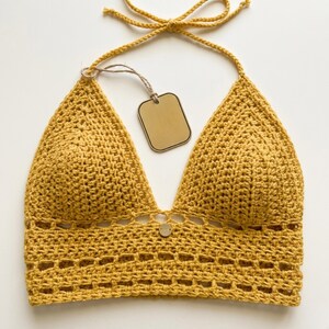 Puede incluir: Top halter de ganchillo amarillo mostaza con cuello en V profundo y bot&oacute;n decorativo. El top presenta una atadura halter y una etiqueta. El dise&ntilde;o de ganchillo incluye un patr&oacute;n calado decorativo en la parte inferior.