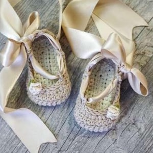 May include: A pair of beige crochet baby shoes featuring a rounded toe and a textured knit pattern. Each shoe is adorned with a small white crocheted flower on the front and includes long, wide champagne-colored satin ribbons for tying.