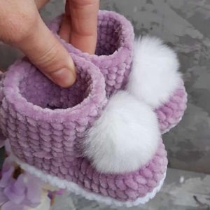 May include: A pair of lavender crochet baby booties made from soft, plush yarn. Each bootie features a thick, textured knit construction and is decorated with a large, fluffy white pom-pom attached to the front, finished with a white trim.