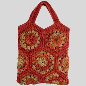 Könnte beinhalten: Eine handgefertigte Häkeltasche in Orange-, Rot- und Beigetönen. Die Tasche hat ein geometrisches Muster aus sechseckigen Formen mit floralen Mittelmuster. Die Tasche hat einen einzigen Griff zum Tragen und besteht aus einem strukturierten Garn.