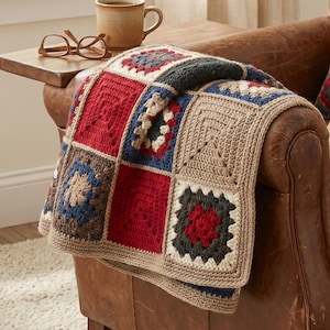 Könnte beinhalten: Eine handgefertigte Häkeldecke, die über einem Ledersessel drapiert ist. Die Decke hat ein Patchwork-Design mit roten, blauen, beigen und grauen Quadraten. Eine braune Tasse und eine Brille liegen auf einem hölzernen Beistelltisch.