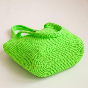 May include: A bright green, crocheted tote bag. The bag features a rounded rectangular base and two handles. The crochet stitches are visible, creating a textured surface. The bag is made of a soft, flexible material.