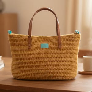 Op de afbeelding: Mosterdgele gehaakte tote bag met bruine leren handvatten en accenten. De tas heeft een rechthoekige vorm, een klein turquoise label en turquoise ritstrekkers. De tas staat op een houten oppervlak.