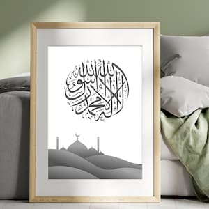 May include: Framed art print featuring Islamic calligraphy in a circular design, rendered in black against a white background. The artwork includes a grayscale illustration of a mosque with domes and minarets. The frame is a light wood color.