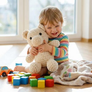 May include: A smiling child hugs a cream-colored crocheted teddy bear. The child wears a rainbow striped shirt and sits on a wooden floor surrounded by colorful wooden blocks and a toy car. A blanket is nearby.