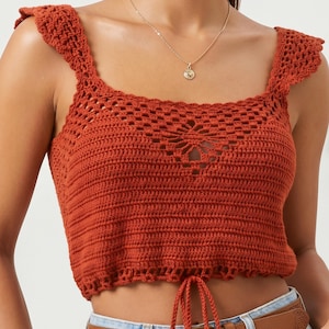 Puede incluir: Top de ganchillo color óxido con cuello cuadrado y tirantes con volantes. El top presenta un cordón en la cintura y un diseño calado floral en la parte delantera. También se ve un collar de oro.