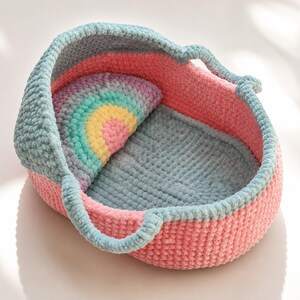 May include: A crocheted bassinet featuring a soft, textured yarn construction. The exterior is pink, the interior base is grey, and the hood is a muted blue. A semi-circular pillow with a rainbow pattern in pastel pink, yellow, and green sits inside.
