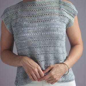 May include: A light gray crochet top with a round neckline and short sleeves. The top features a detailed openwork pattern throughout the bodice and sleeves. The garment is worn over a white undershirt.