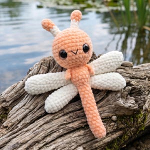 May include: A handmade crochet dragonfly toy in peach and white. The dragonfly has large, glittery eyes and is posed on a piece of driftwood. The wings are white, and the body is peach-colored. The dragonfly is a whimsical, soft toy.