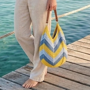 May include: A handmade crochet bag with a chevron pattern in yellow, blue, and white. The bag has brown leather handles and gold-colored hardware. The bag is held by a person standing on a wooden dock near the water.