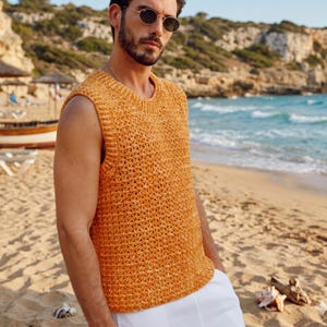 May include: A sleeveless, open-weave, orange-yellow knit top. The top has a crew neck and a textured appearance. The person is wearing sunglasses and white pants. The background is a beach with sand, ocean, and cliffs.
