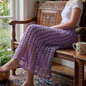 Puede incluir: Una falda de ganchillo lavanda con un dobladillo festoneado. La falda se usa mientras está sentado en un banco de madera. El banco tiene detalles florales tallados. Una taza blanca con ilustraciones de gatos está sobre una mesita auxiliar.