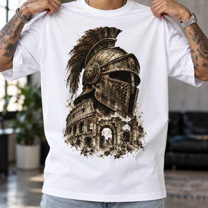 May include: A white t-shirt featuring a detailed graphic print of a weathered Roman gladiator helmet with a crest, positioned above an illustration of the Colosseum. The artwork uses shades of brown, bronze, and black to create a textured effect.