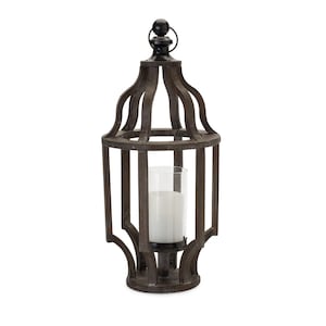 May include: A decorative, dark brown metal lantern with a clear glass cylinder inside, holding a white candle. The lantern has an arched top and a black finial. The design is reminiscent of a birdcage.