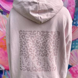 Könnte beinhalten: Hellrosa Kapuzenpullover mit Leopardenmuster auf dem Rücken. Das Wort "MOM" ist in der Mitte des Musters aufgedruckt. Der Hoodie hat eine Kapuze und lange Ärmel.