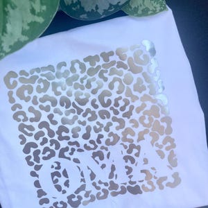 Könnte beinhalten: Weißes quadratisches Kissen mit goldenem Leopardenmuster und dem Wort "OMA" in großen Buchstaben. Das Kissen besteht aus einem weichen Stoff und eignet sich für die Inneneinrichtung.