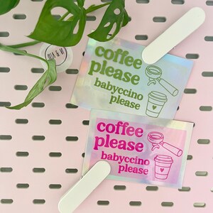 Könnte beinhalten: Zwei holografische Aufkleber mit dem Text "coffee please babyccino please" in Grün und Pink, zusammen mit Kaffee-Illustrationen. Jeder Aufkleber wird von einer weißen Klammer gehalten. Der Hintergrund ist eine rosa Lochplatte.