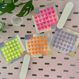 Könnte beinhalten: Vier quadratische Stickerbögen mit Herzmustern in Neonrosa, Orange, Gelb und Lila. Ein weißer Applikator befindet sich neben den Aufklebern. Die Aufkleber befinden sich auf einer rosa Oberfläche.