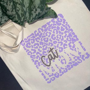 Könnte beinhalten: Cremefarbene Tragetasche mit lila Leopardenmuster und dem Schriftzug "Cat Mom" in Glitzer. Die Tasche hat cremefarbene Griffe und ist mit einer grünen Pflanze verziert.