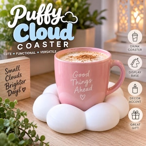 May include: A pink ceramic mug with the white text Good Things Ahead and a small heart icon, resting on a white, cloud-shaped coaster. The coaster has a rounded, multi-lobed design. The mug contains a beverage topped with brown cinnamon powder.