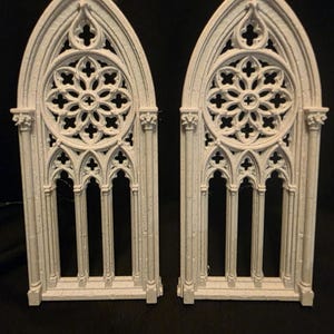 May include: Two off-white, arched window frames featuring intricate, openwork tracery and a central rose window design. Each frame displays vertical columns, pointed arches, and detailed stone-like textures, mimicking architectural elements.