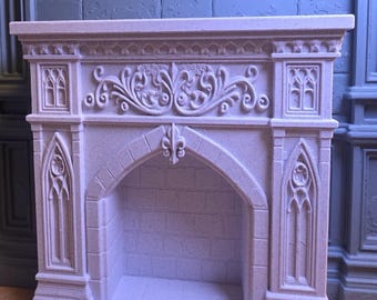 May include: A light purple miniature fireplace mantel featuring intricate carved details, including a central fleur-de-lis, arched side panels with gothic window motifs, and a textured brick-patterned interior firebox with a flat top shelf.