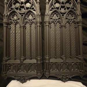 May include: Two identical black decorative panels featuring a detailed gothic arch window design. Each panel displays intricate vertical columns, a lattice pattern, and a quatrefoil motif at the top, all rendered in a matte black finish.