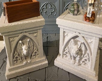 May include: Two off-white speckled stone-like pedestals featuring carved gargoyles within arched alcoves. One pedestal holds a brown rectangular wooden box, while the other supports a clear glass jar with a label reading Hogwarts.