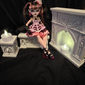 May include: A doll with black and pink hair, wearing a pink and black dress with a spiderweb pattern, sits on a stone-like bench next to a miniature fireplace and a small pillar, both featuring LED tea lights with a warm glow.