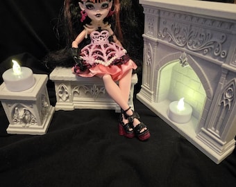 May include: A doll with black and pink hair, wearing a pink and black dress with a spiderweb pattern, sits on a stone-like bench next to a miniature fireplace and a small pillar, both featuring LED tea lights with a warm glow.