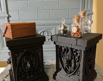 May include: Two black square pedestals with gothic bat carvings on the sides. A brown wooden box sits on the left pedestal. On the right pedestal are three small glass bottles with cork stoppers, one containing small red items and a label.