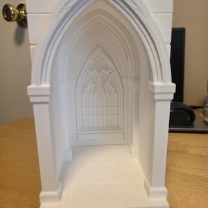 May include: A white 3D printed architectural model of a gothic cathedral interior featuring a pointed arch entryway, a detailed traceried window at the back, and a tiled floor. The structure is composed of smooth, matte white material.