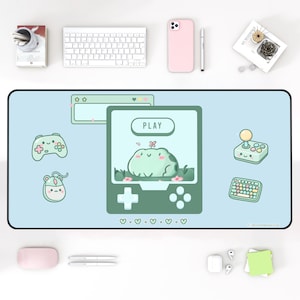 May include: A rectangular desk mat with a light blue background featuring a central green handheld game console graphic with a frog character on the screen and the text PLAY. Surrounding graphics include a game controller, mouse, joystick, and keyboard.