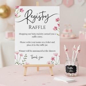 May include: A white sign on a wooden easel with the words "Registry RAFFLE" in black script. The sign is decorated with pink floral accents. A glass jar labeled "Raffle Jar" holds pens with flower toppers.