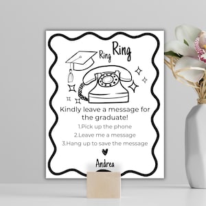 以下が含まれることがあります： 電話と卒業帽子のイラストが描かれた白黒のサインで、「Ring Ring」の文字が入っています。サインには「卒業生にメッセージを残してください！」と指示と名前の「Andrea」が書かれています。