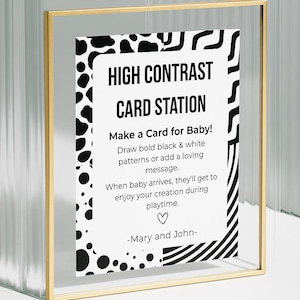 May include: A framed sign with gold trim, featuring the text "HIGH CONTRAST CARD STATION." The sign encourages creating cards with black and white patterns or a loving message for a baby. The sign is a baby shower or nursery decor item.