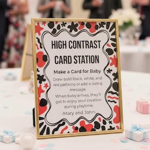 Puede incluir: Un letrero rectangular con marco dorado, con el texto "HIGH CONTRAST CARD STATION". El letrero anima a los invitados a crear tarjetas para un bebé, con patrones en negro, blanco y rojo. El letrero está decorado con formas negras, rojas y blancas.