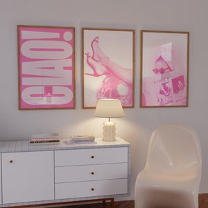May include: Three framed pink art prints. The left print says "CIAO!" in large white letters. The middle print shows a person's legs and feet in pink high heels. The right print features a Vogue magazine cover. A white lamp and cabinet are in the foreground.