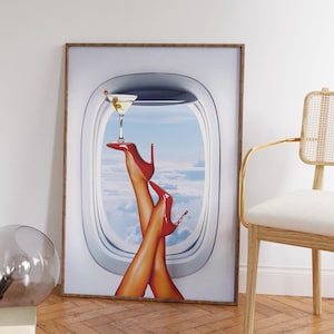 Pode incluir: Uma impressão emoldurada de pernas de mulher em saltos altos vermelhos saindo de uma janela de avião com uma taça de martini apoiada em um de seus pés. O fundo é um céu azul com nuvens brancas.