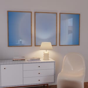 May include: A white dresser with three drawers and gold knobs sits on a wood floor. A white chair with a curved back sits to the right of the dresser. A lamp with a white shade sits on the dresser. Three framed pictures with blue backgrounds hang on the wall above the dresser.