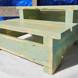 Peut inclure: Un tabouret en bois à deux marches peint en vert clair avec une finition vieillie. Le tabouret a un design simple avec un dessus rectangulaire et une marche inférieure.