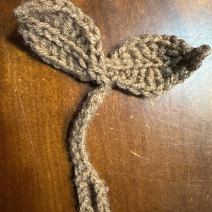 May include: A brown crochet sprout featuring two textured leaves attached to a single stem. The yarn work displays a visible stitch pattern throughout the entire piece, creating a soft, flexible, and lightweight plant-like shape in a solid earth tone.