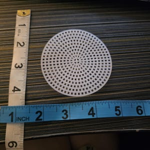 Peut inclure: Une toile en plastique blanche et circulaire avec un motif quadrillé, d'environ 10 cm de diamètre. Un ruban à mesurer bleu et blanc est à côté de la toile, indiquant la mesure en pouces. La toile est sur une surface sombre et texturée.
