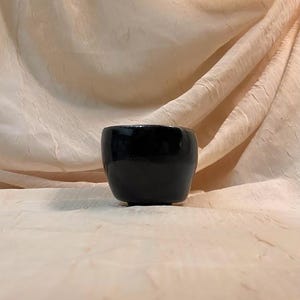 Op de afbeelding: Een zwarte keramische pot met een gladde, glanzende afwerking. De pot staat op een lichtbeige stoffen oppervlak.