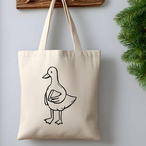 A beige canvas tote bag hanging on a wall next to festive evergreen décor. The bag displays a hand-drawn goose holding a book, printed in simple black line art. The mockup highlights how the bookish goose SVG works beautifully on tote bags, bookstore merchandise, reusable shopping bags, and cozy aesthetic gifts for readers. Clean lines make the design suitable for screen printing, heat transfer vinyl, and sublimation projects.